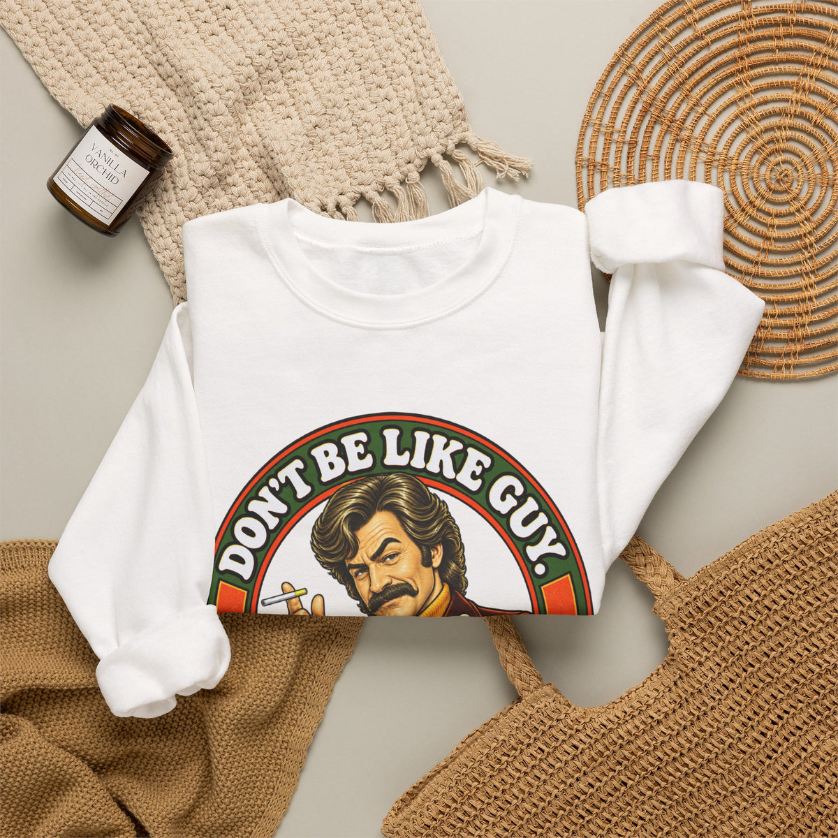 Flat lay of white crewneck with Don’t Be Like Guy graphic and cozy home props